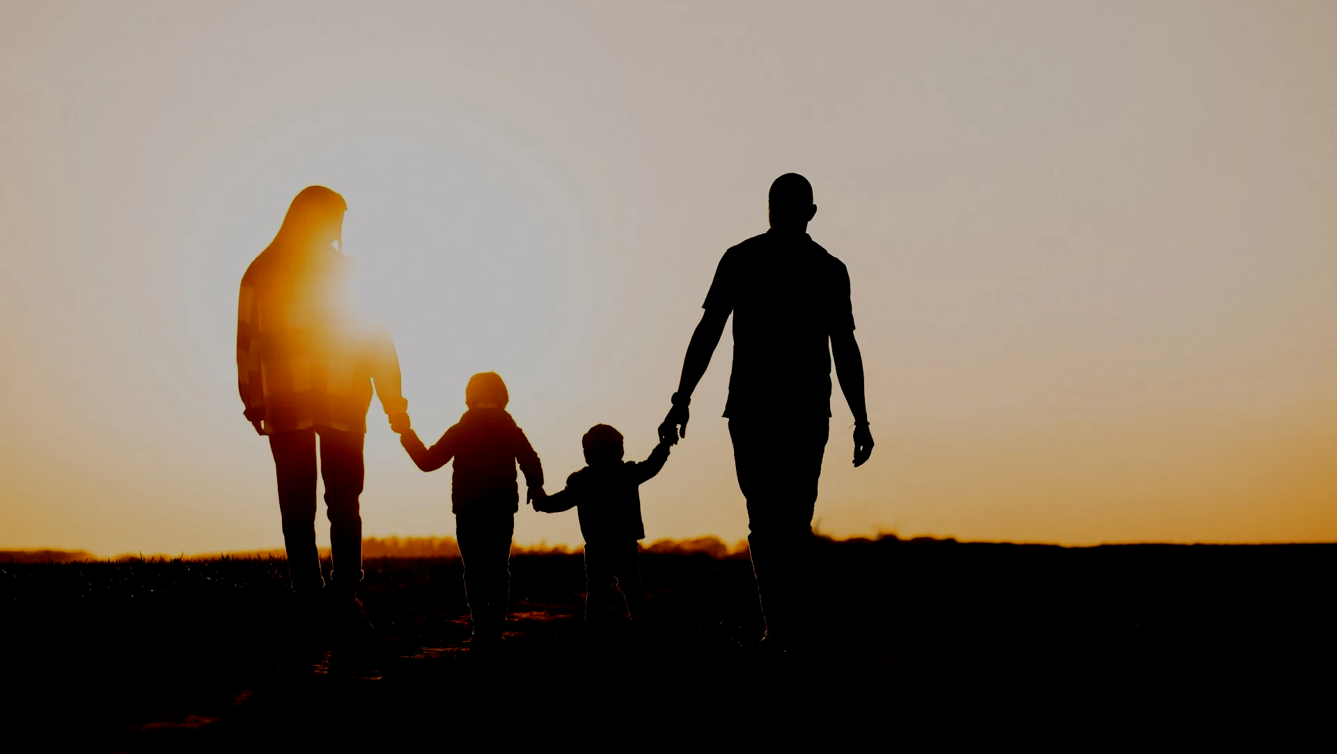 Family of four holding hands, walking towards sunset. Silhouettes against a bright orange sky.