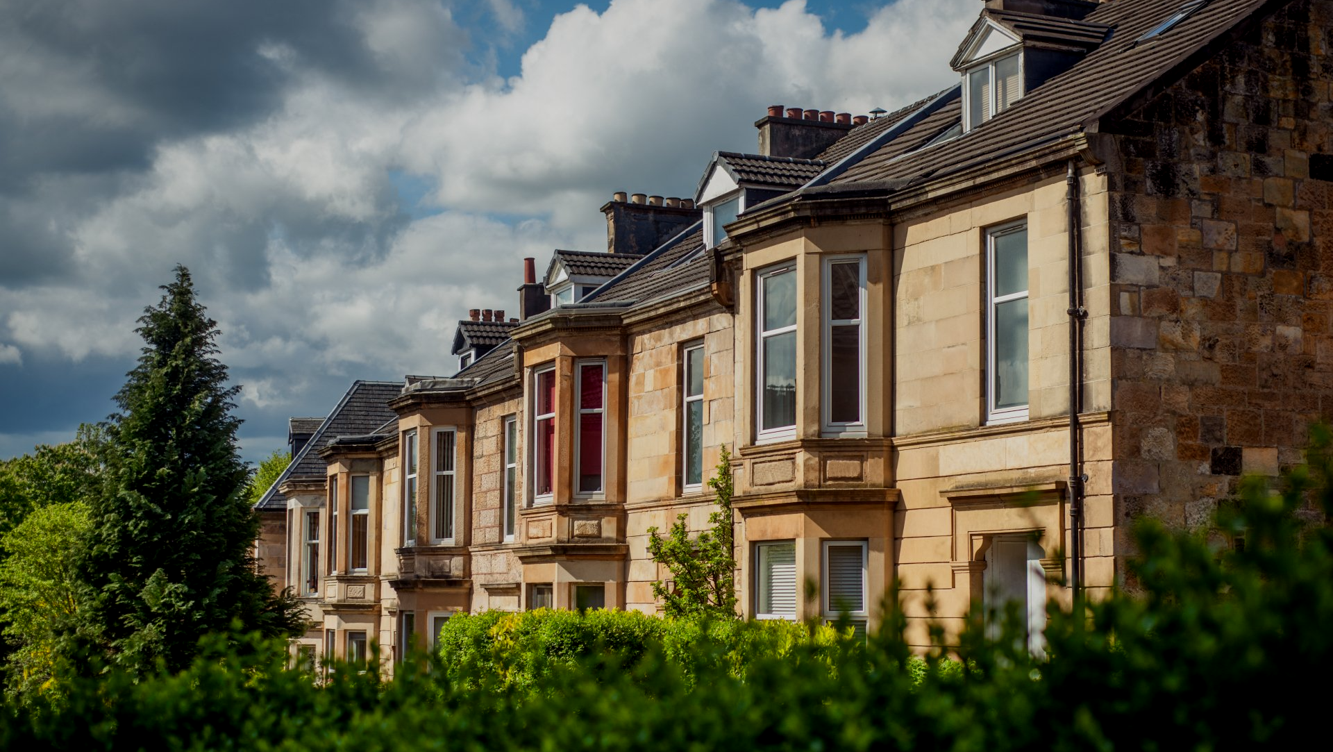 Row of sandstone houses with bay windows, under a cloudy sky, partly obscured by green trees.