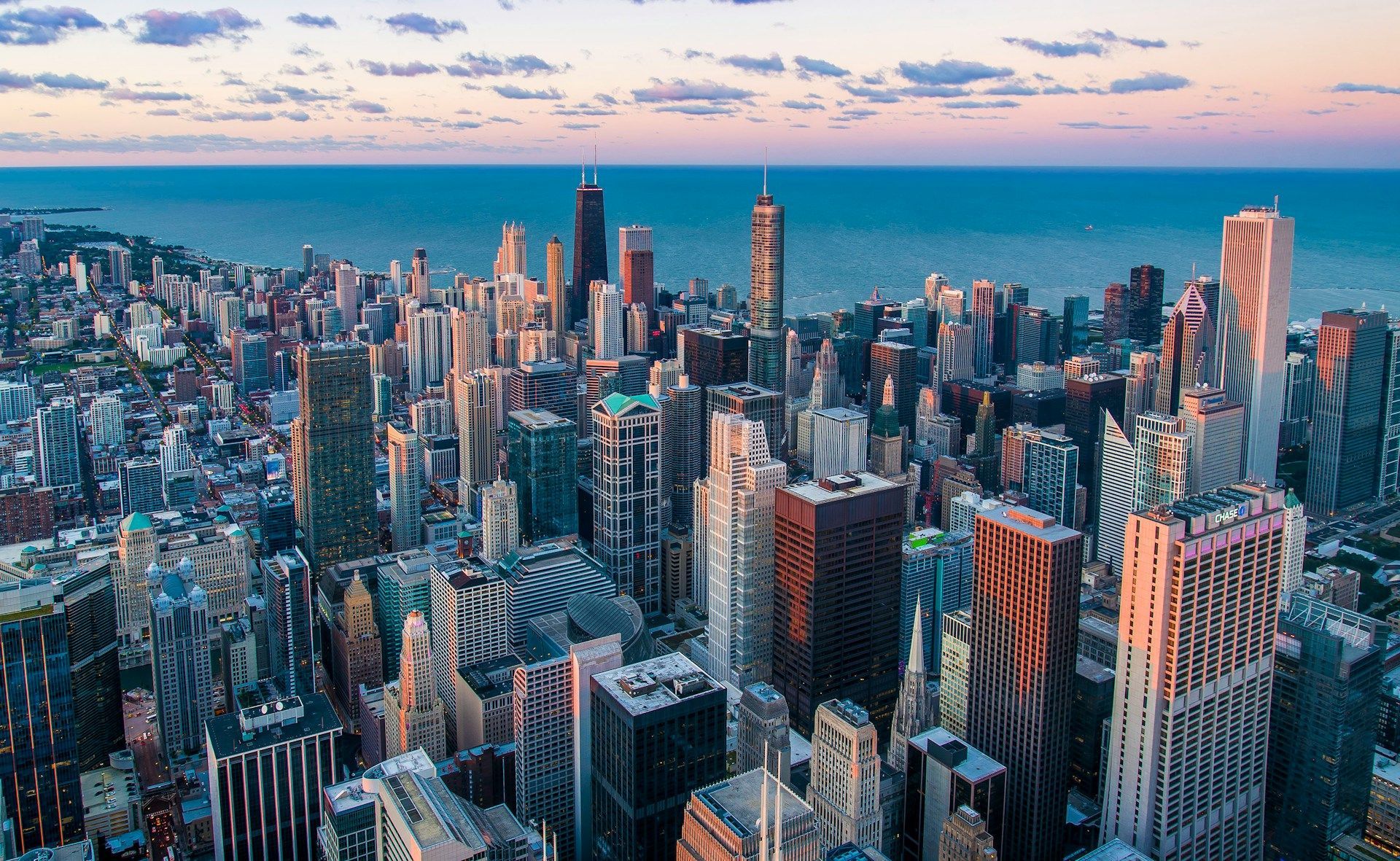 Aerial view of a dense city skyline at sunset, with tall skyscrapers beside a blue body of water.