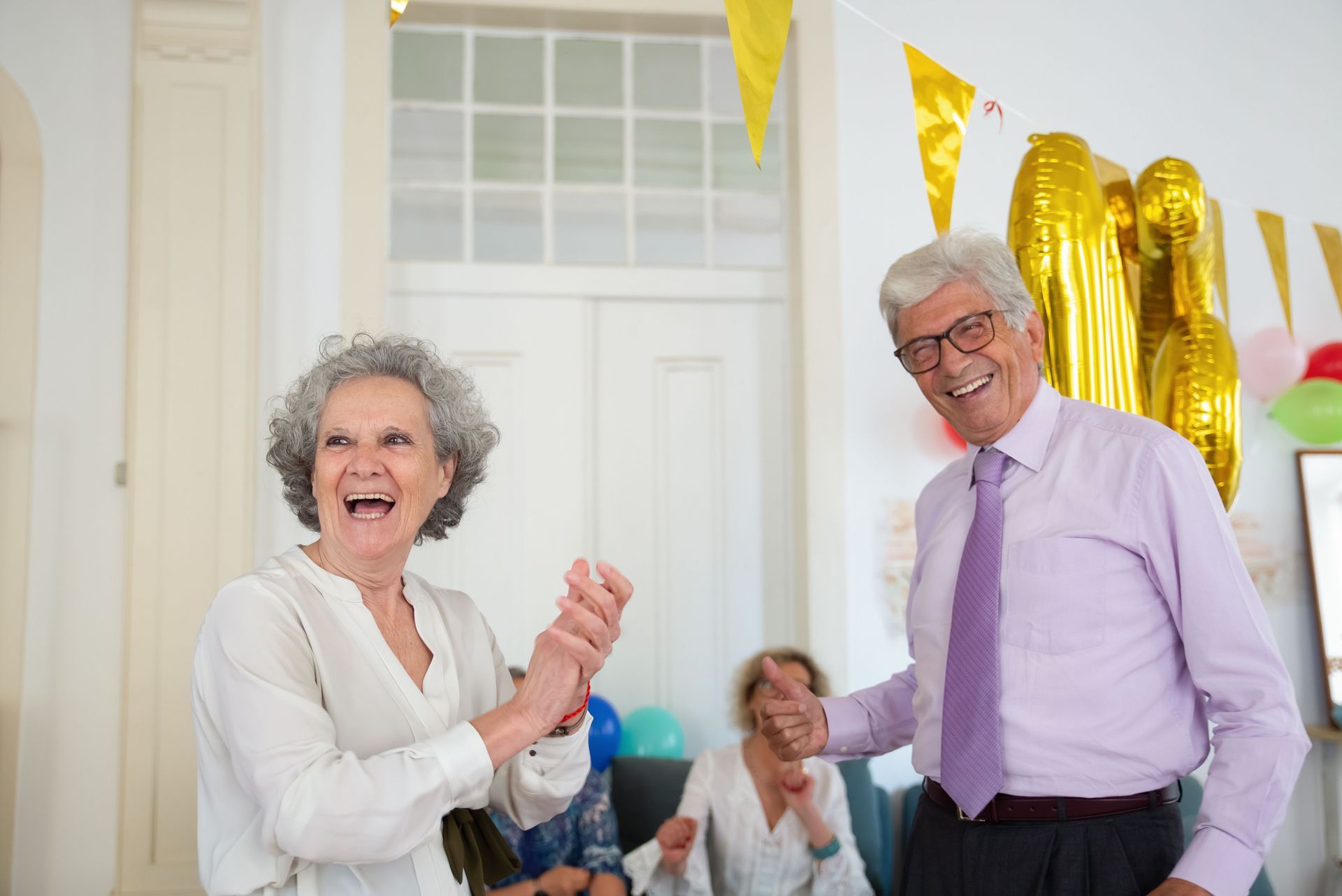 Two smiling people in party clothes celebrate indoors with gold balloons and seated guests in the background