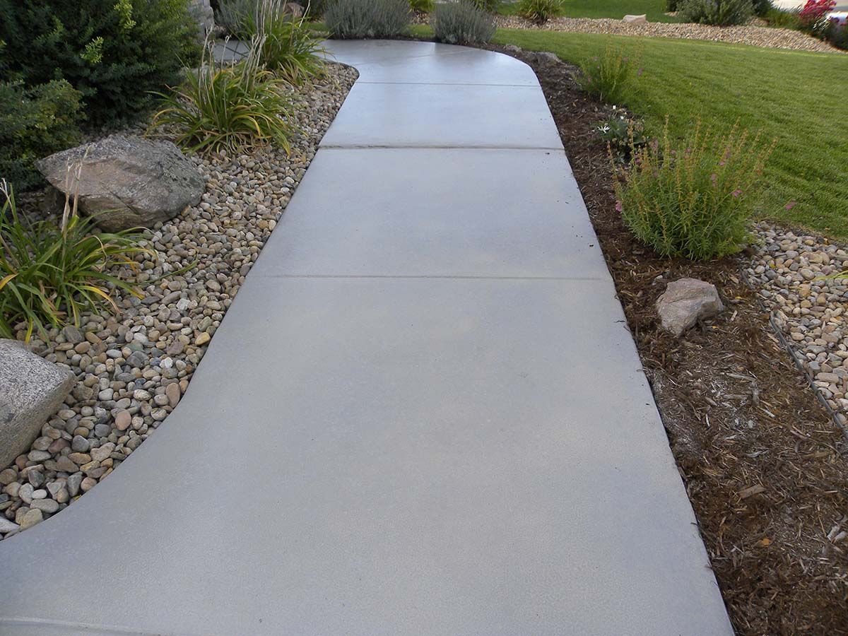 Gray concrete walkway bordered by plants, rocks, and grass.