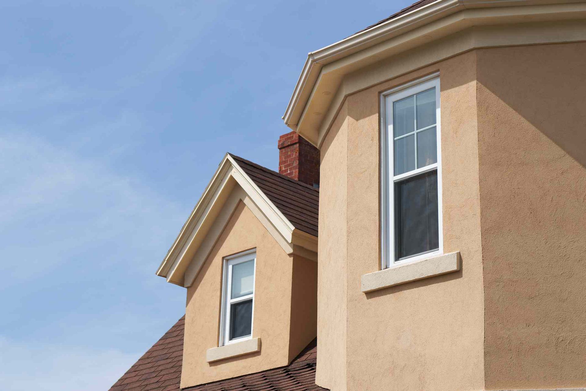 Tan stucco house with white-framed windows, brown roof, and brick chimney against a blue sky.