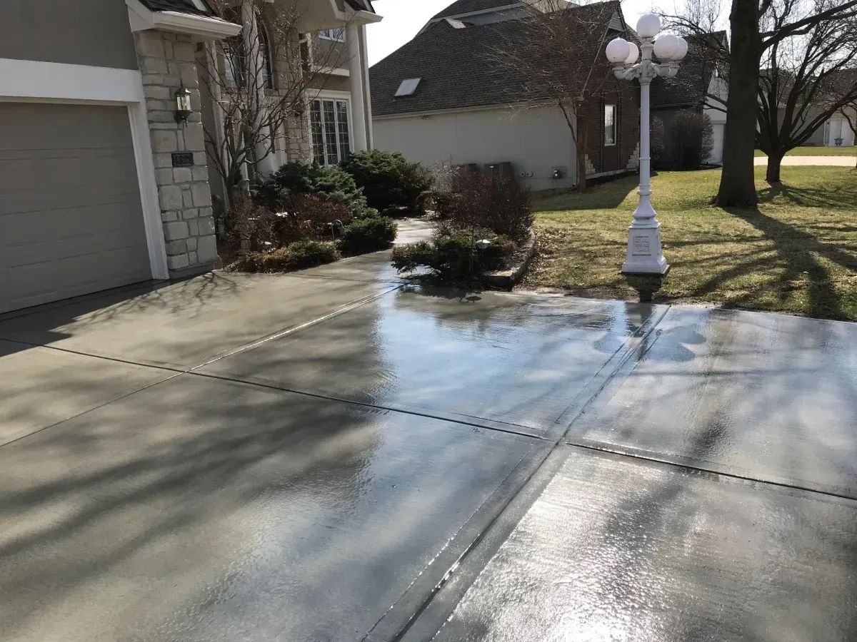 Wet, light gray concrete driveway in front of a house, reflecting sunlight.