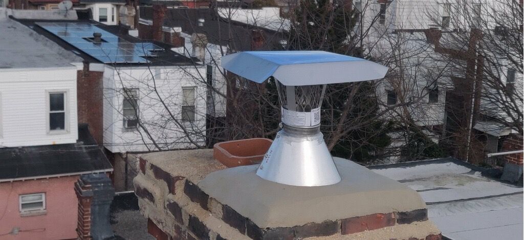 Chimney with metal cap on a brick structure against a backdrop of rooftops and bare trees.