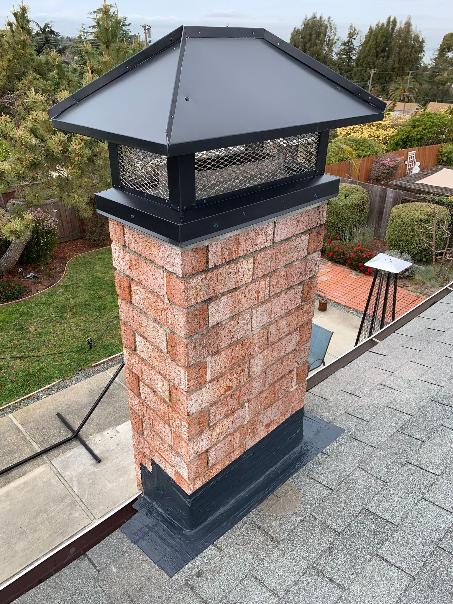 Brick chimney with black cap on a rooftop, surrounded by greenery.