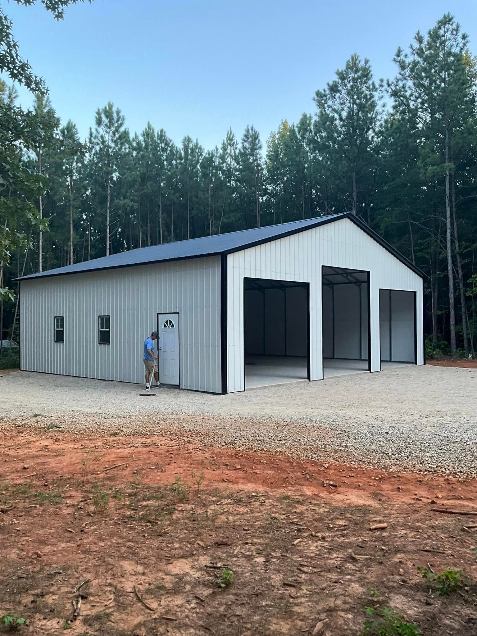 Gray metal workshop with open bays and a person standing outside, surrounded by gravel and trees