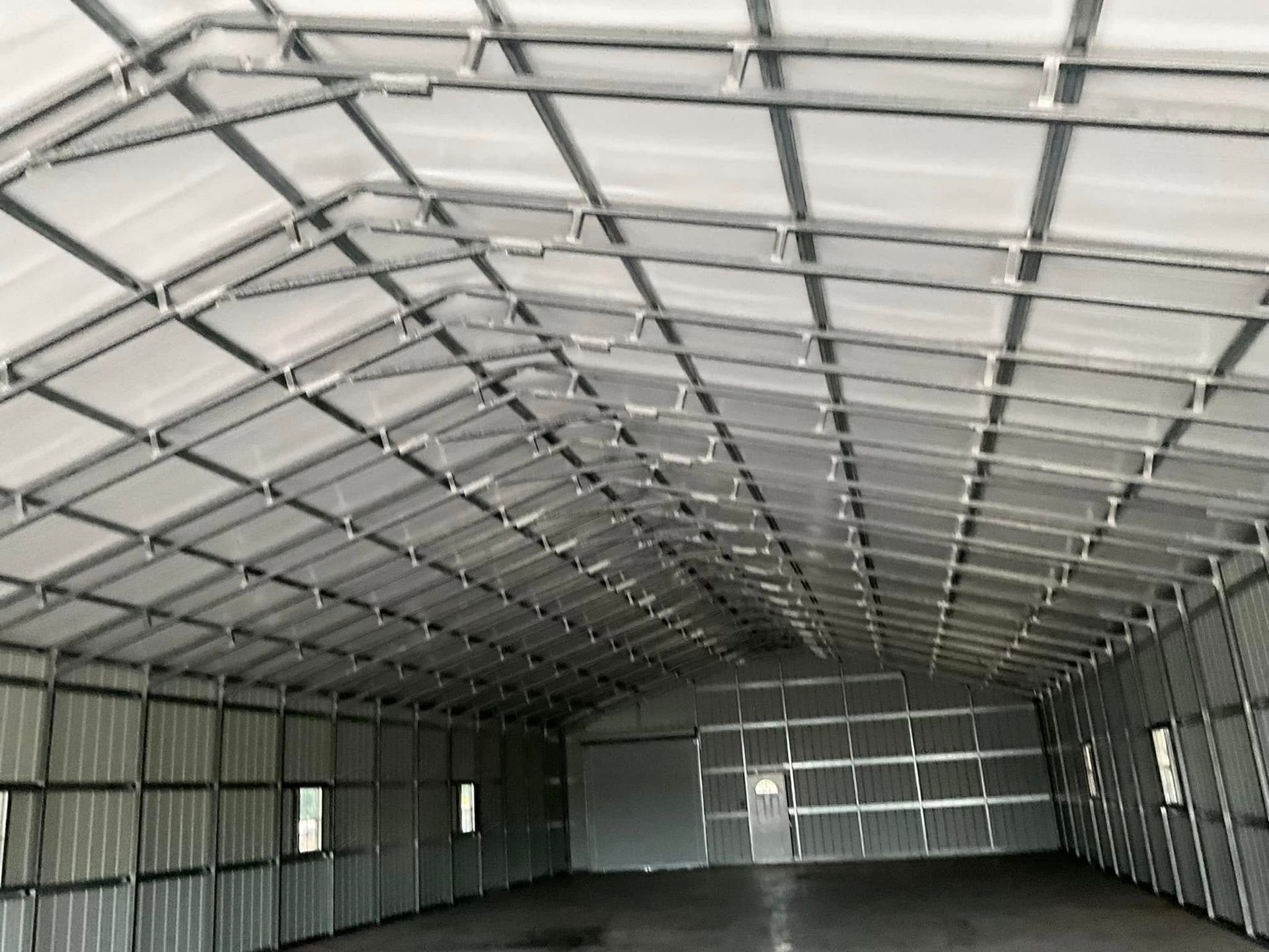 Interior of an empty metal warehouse with a corrugated roof and walls, viewed from one end toward large closed doors.