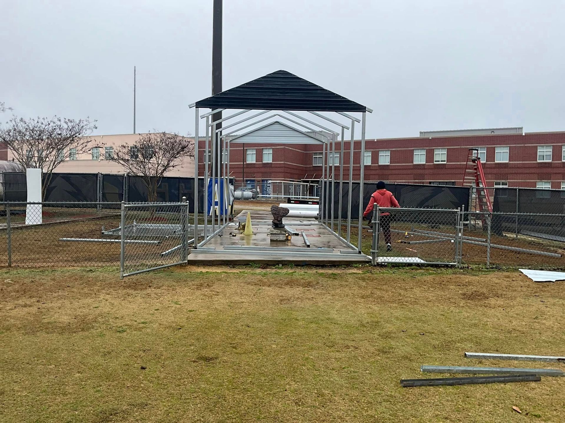Outdoor pavilion under construction with workers, concrete slab, and a brick building in the background.