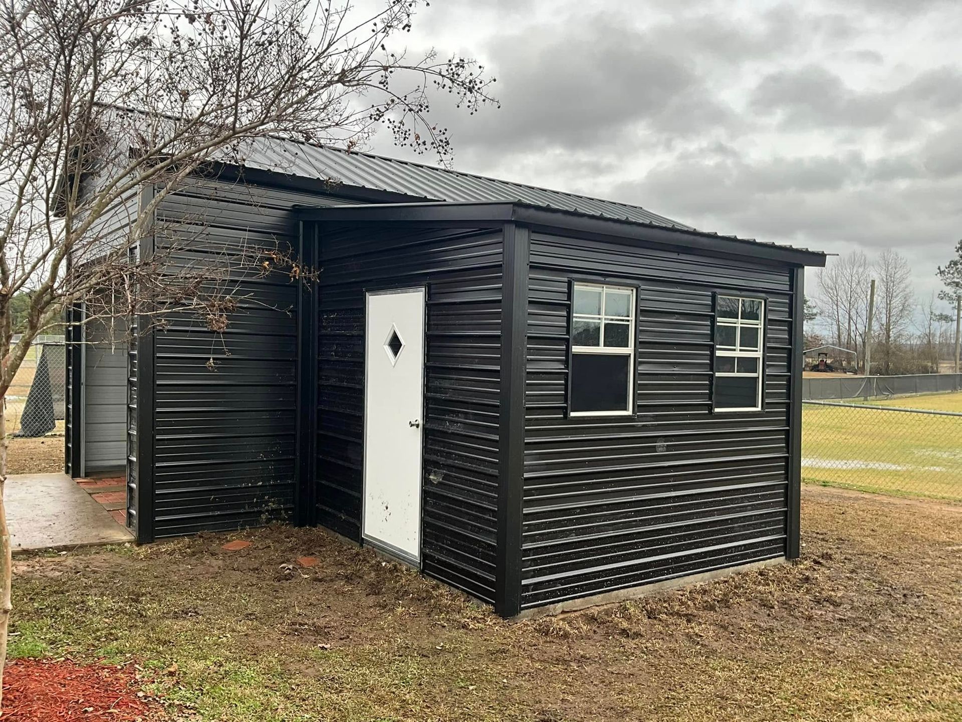 Small black shed with white door and windows in a grassy field under a cloudy sky