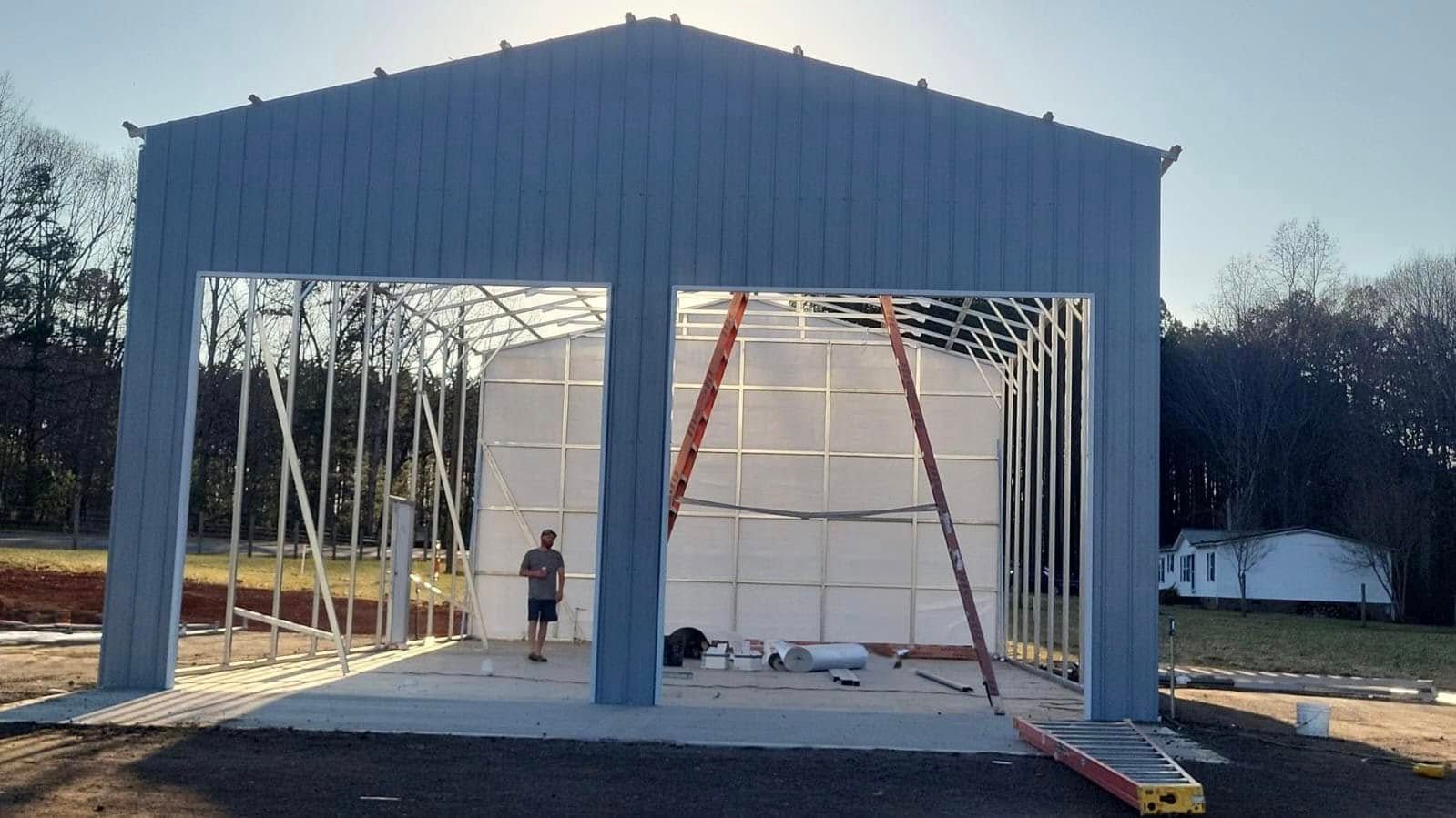 Partially built metal shed frame with white wall panels and ladders in a yard at dusk