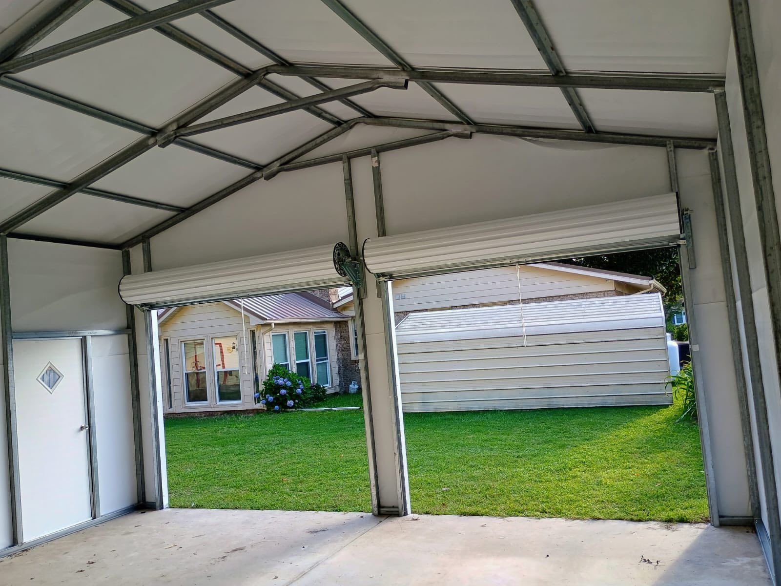 Inside a carport garage, open roll-up doors frame a green yard and a small house in the background.