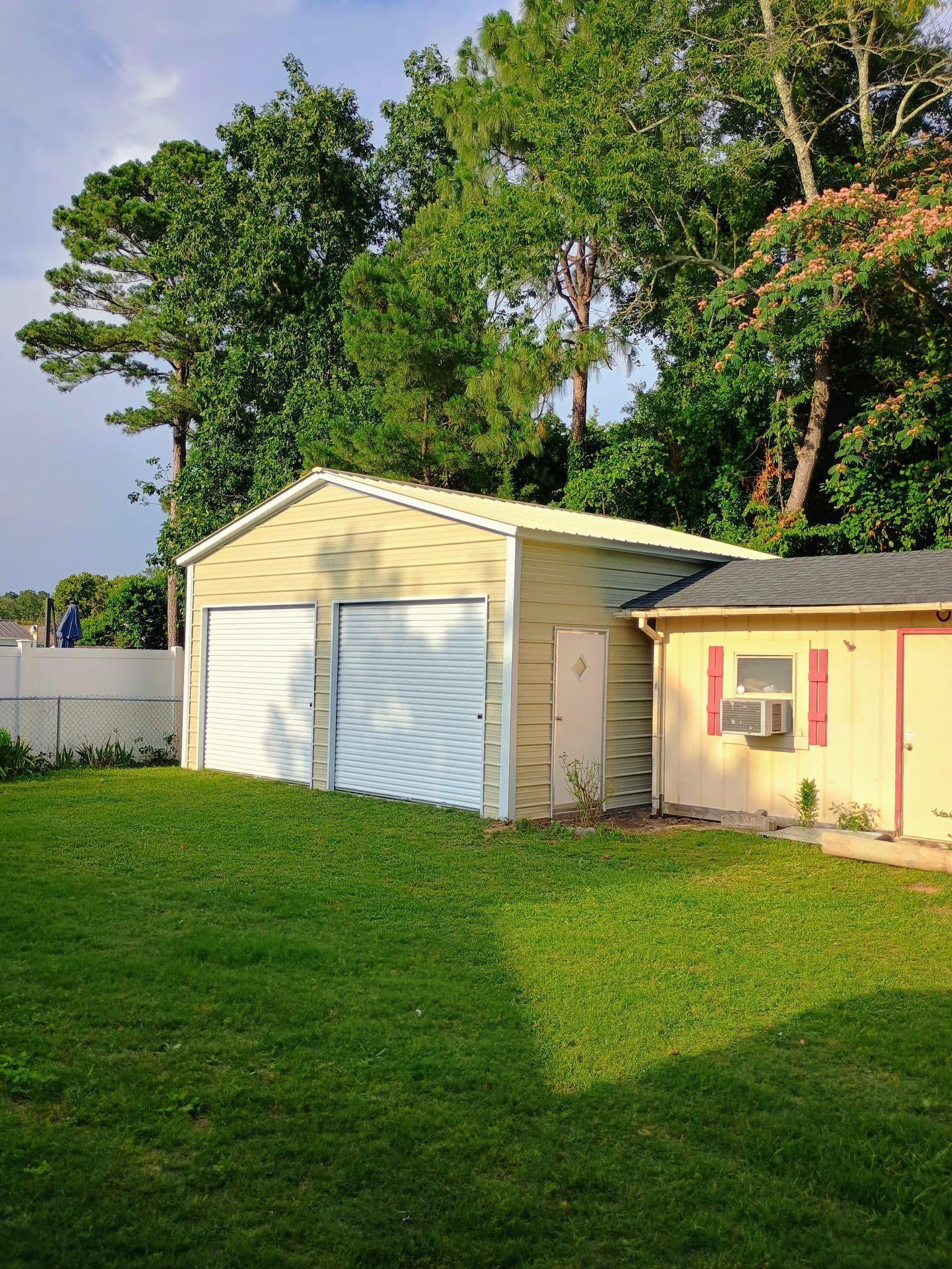 Backyard with a beige garage, small shed, and large green trees in warm sunlight