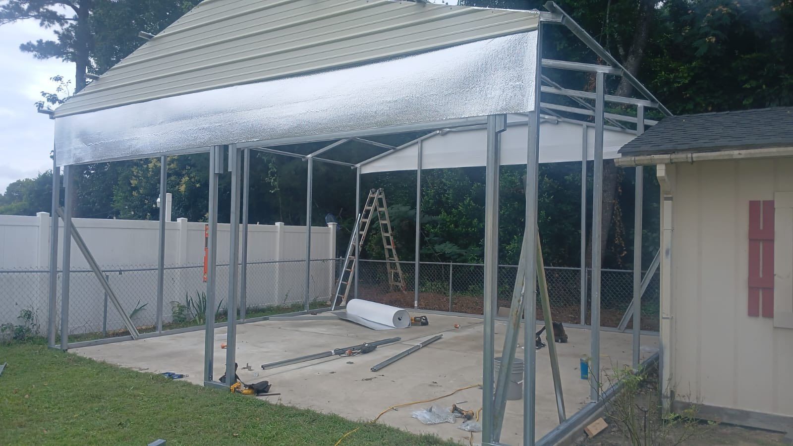 Partially built metal carport frame with corrugated roof panels beside a small building and ladder