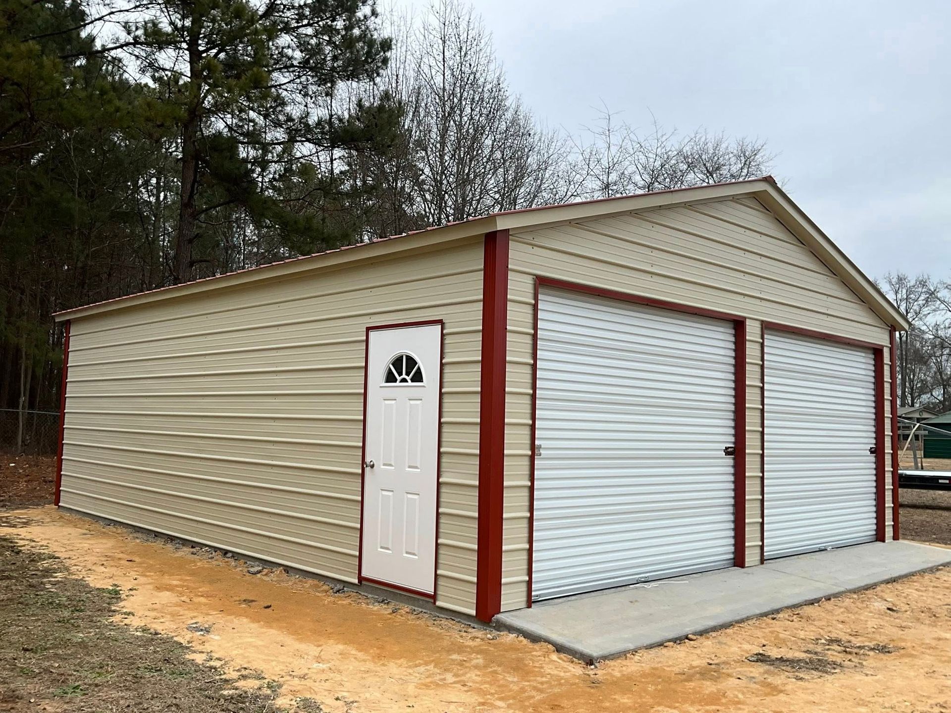 Tan metal garage with two roll-up doors, white side door, and red trim on a concrete pad in a wooded area
