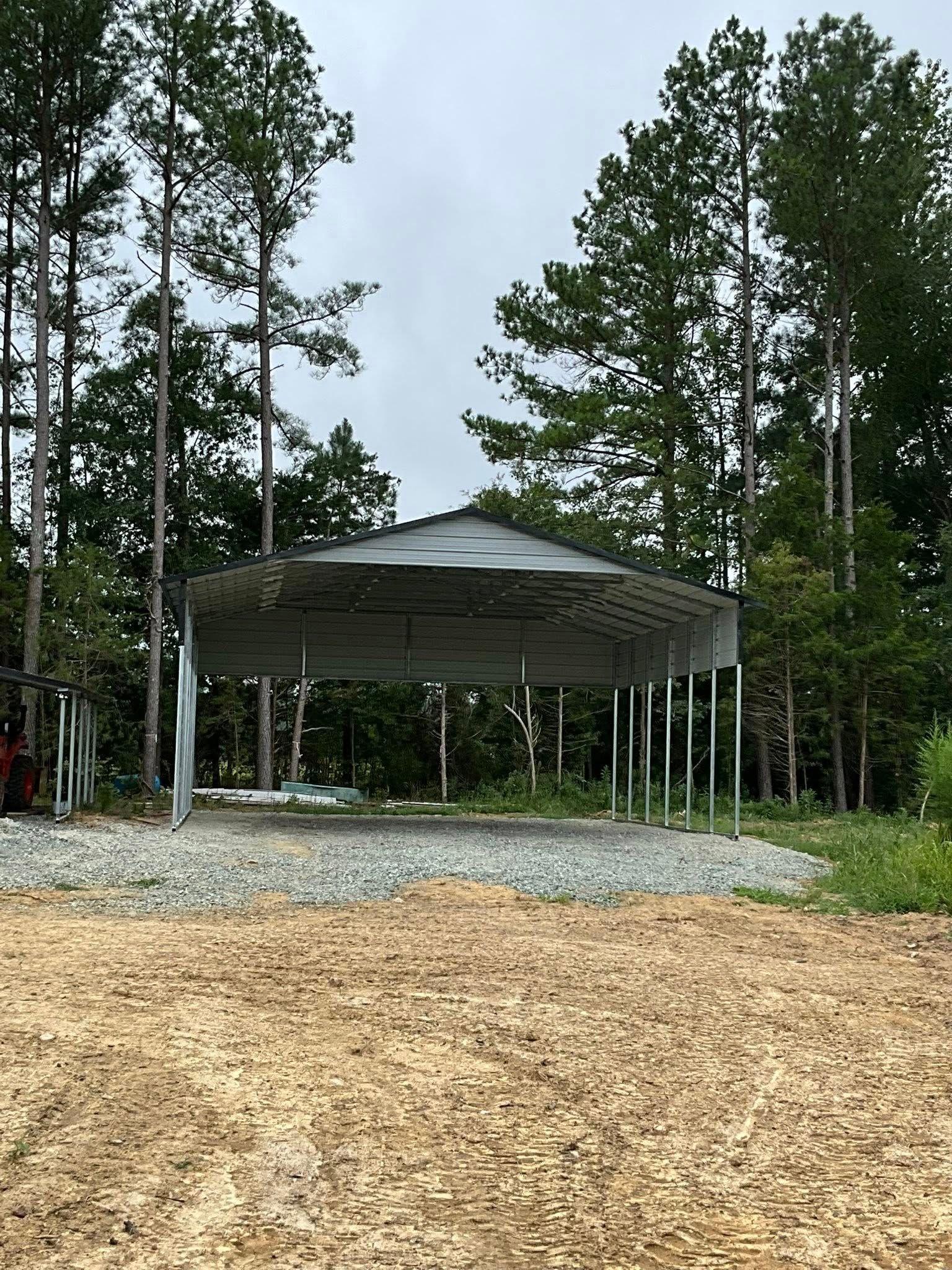 Open-sided metal pavilion in a wooded park with gravel ground and pine trees.