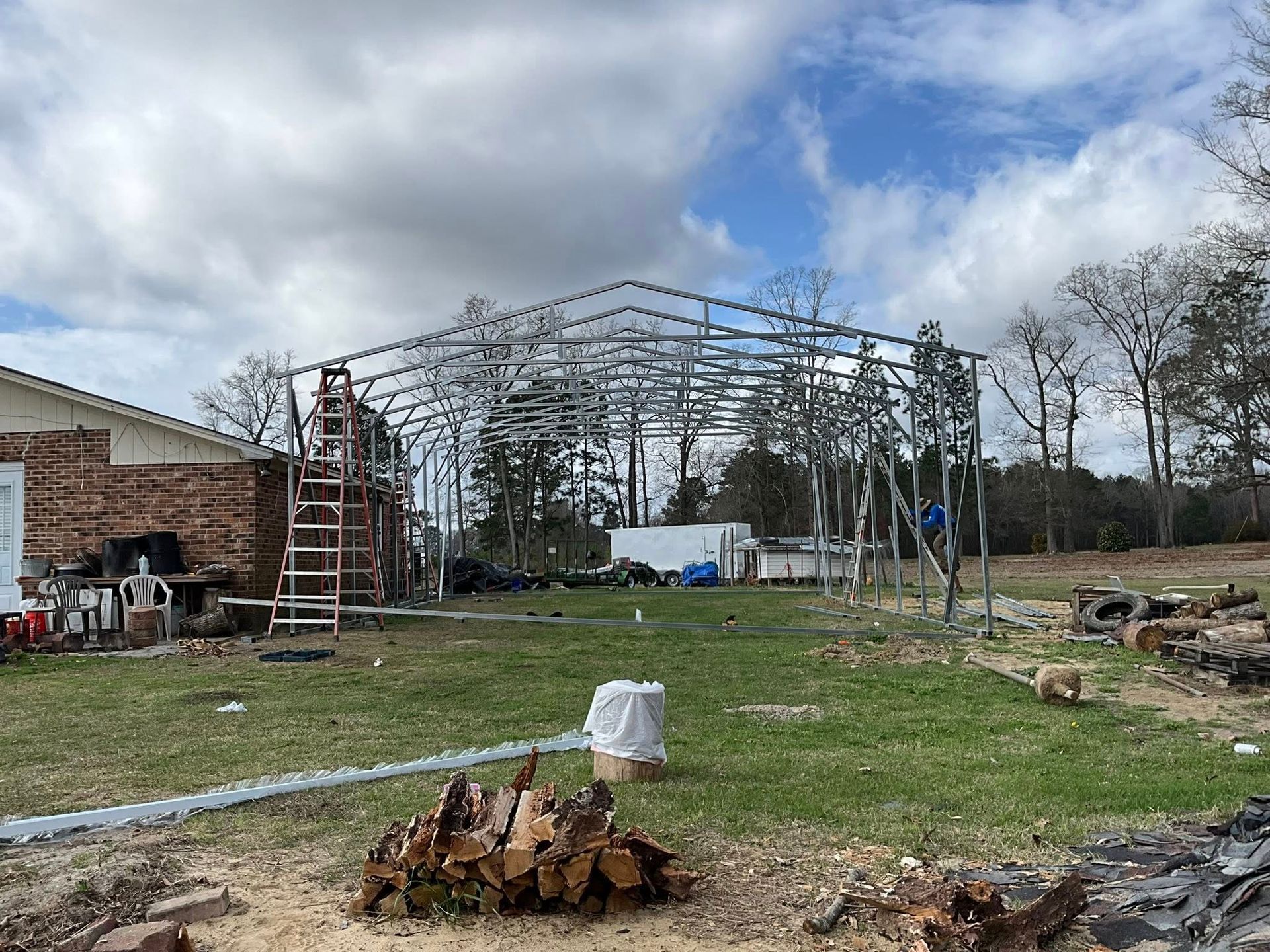 Metal framework under construction in a grassy yard, with a ladder, trees, and cloudy sky.