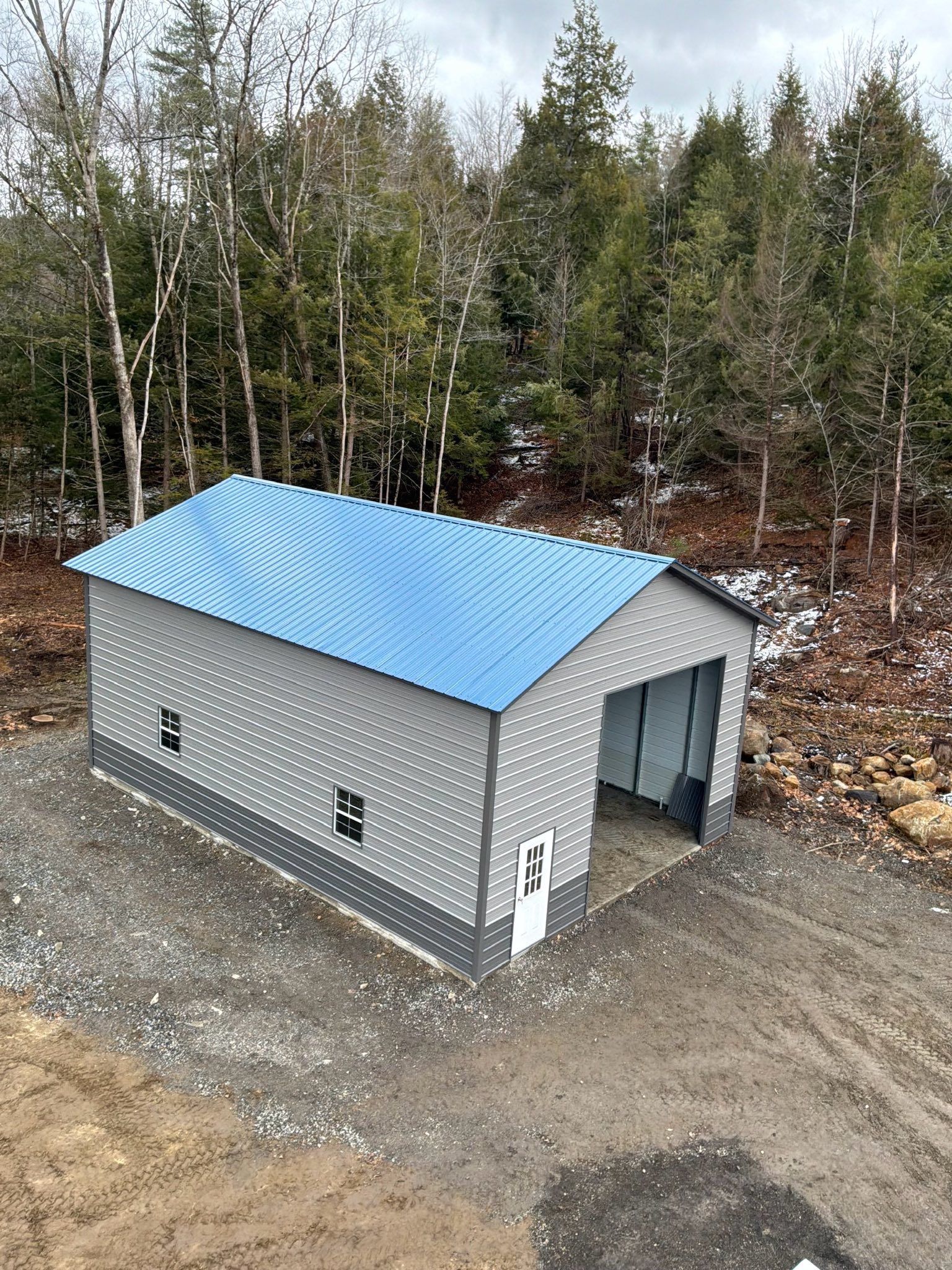 Small gray metal shed with a blue roof on a gravel lot beside a wooded area.