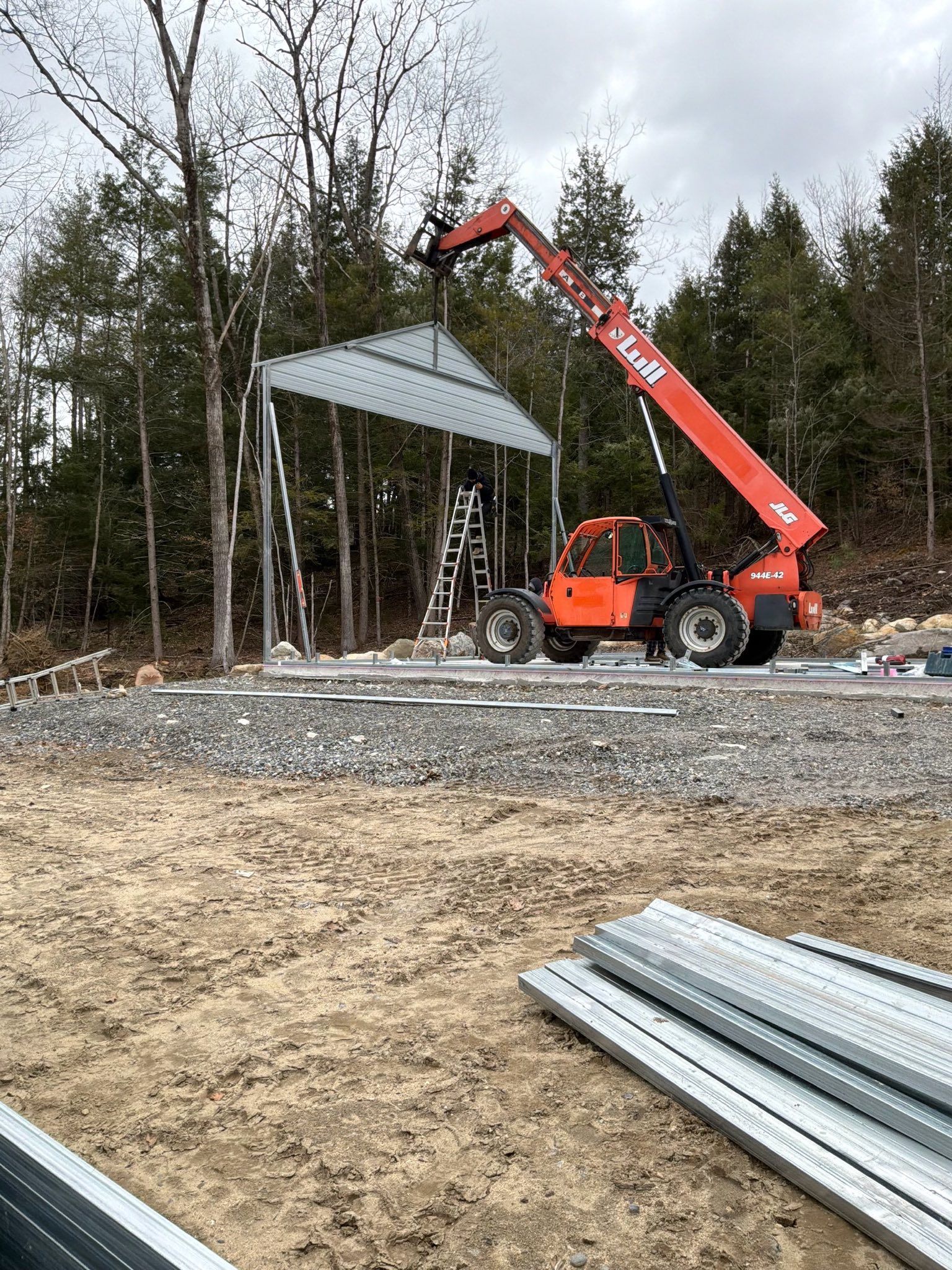 Orange crane lifting a metal roof frame at a construction site with gravel and stacked steel in foreground