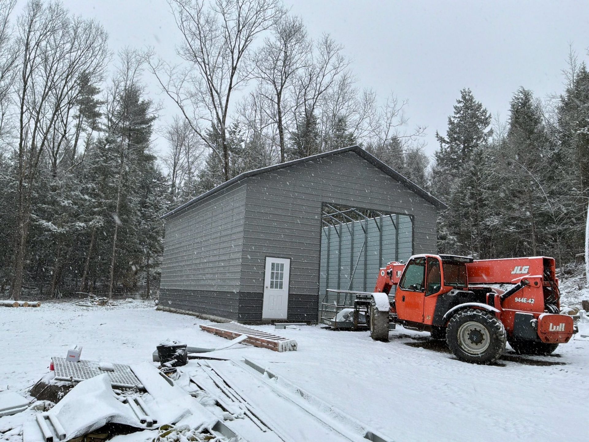 Snowy yard with a gray metal shed and an orange forklift beside construction materials.
