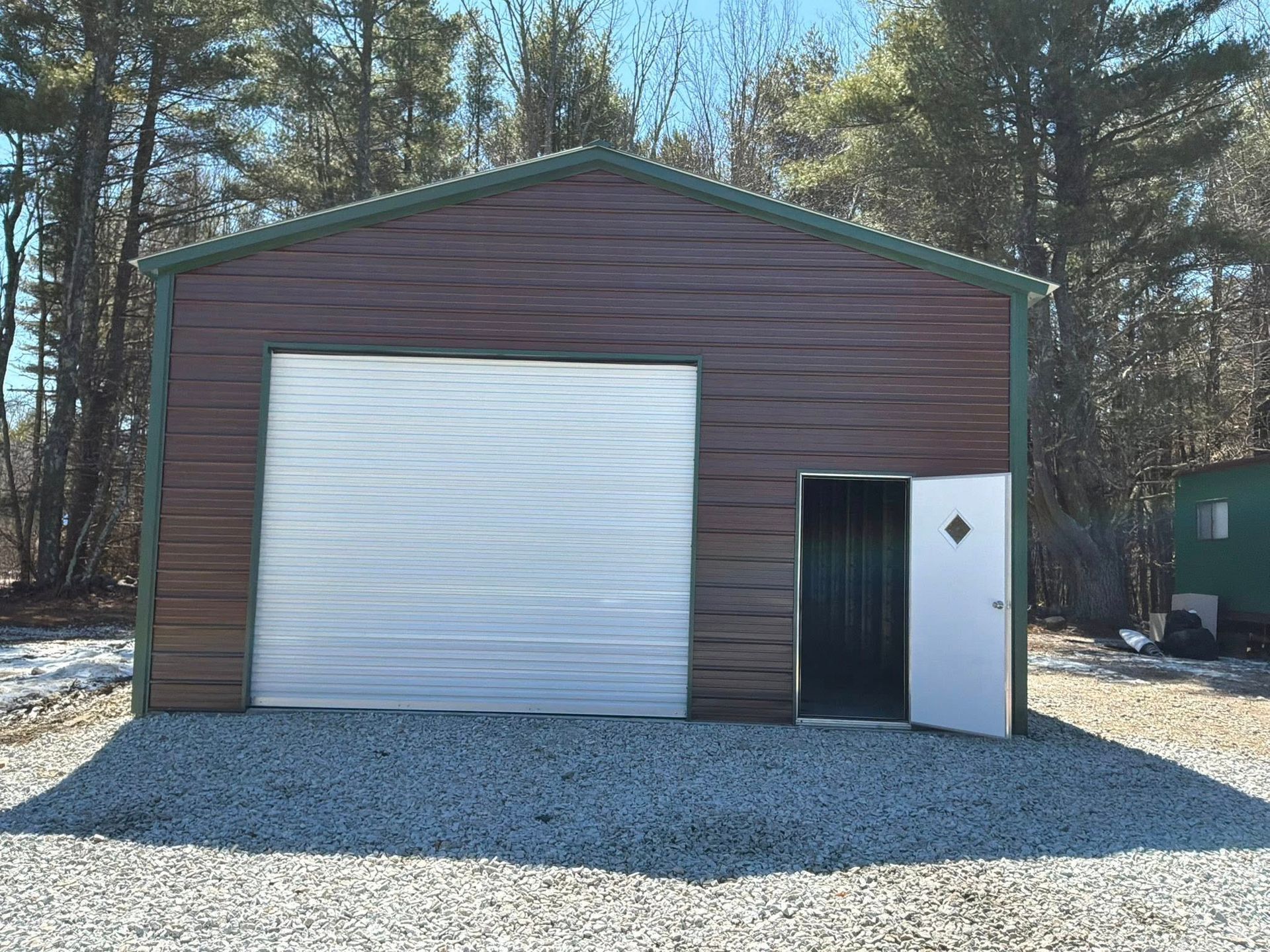 Brown metal garage with a white roll-up door and side entry door on a gravel lot in a wooded area