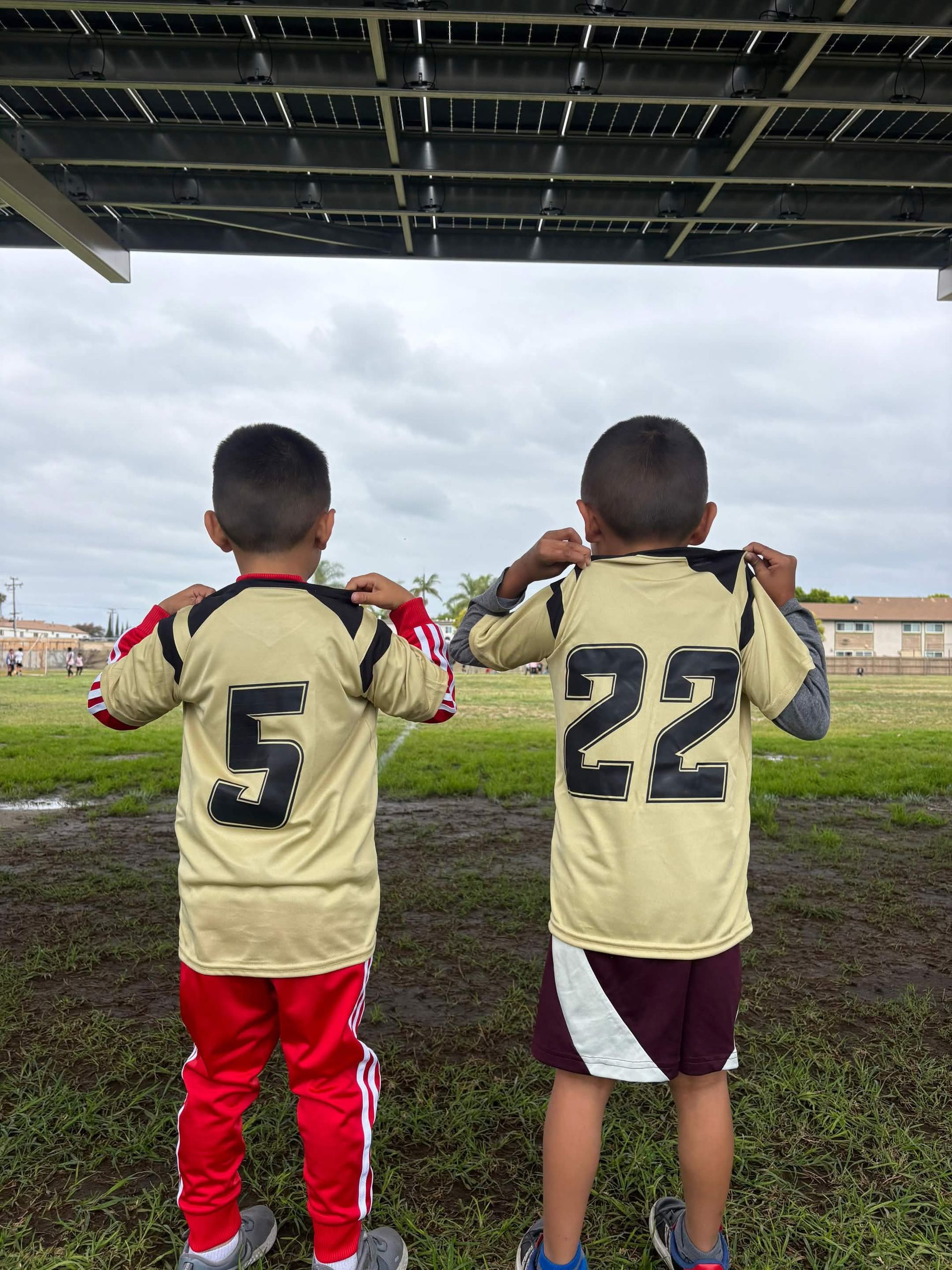 Two children in sports jerseys stand under a roof, facing a field and adjusting their collars.