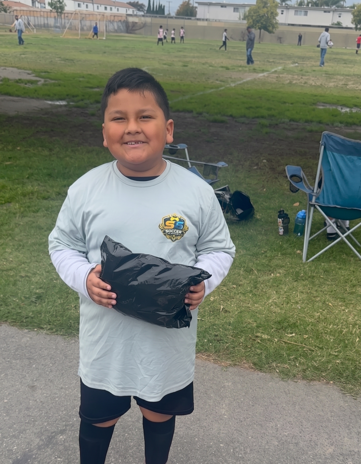 Smiling child in a gray shirt holding a black bag on a park path, with a grassy field behind him