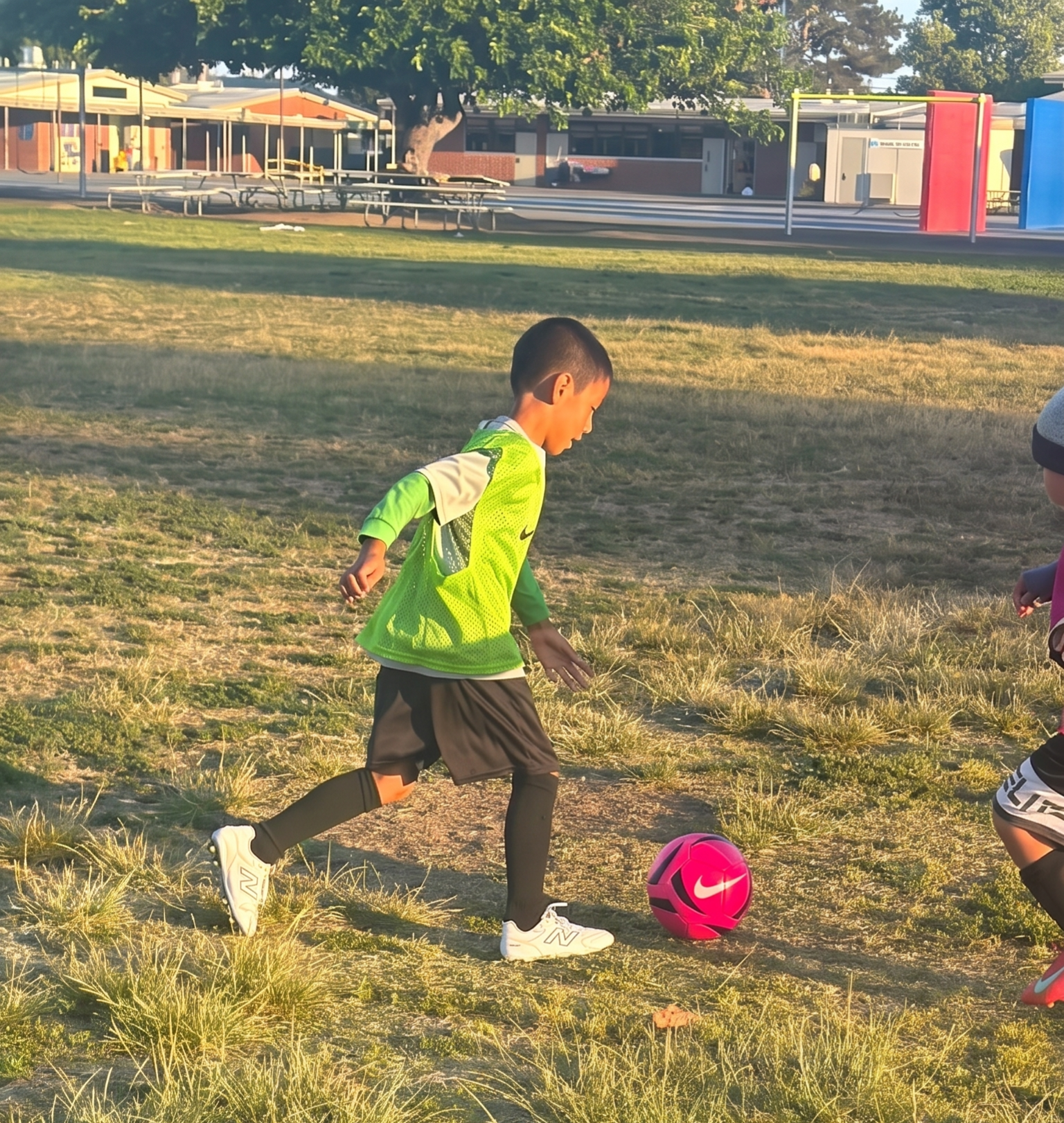 Child in a neon green jacket dribbles a pink soccer ball on a grassy field at sunset.