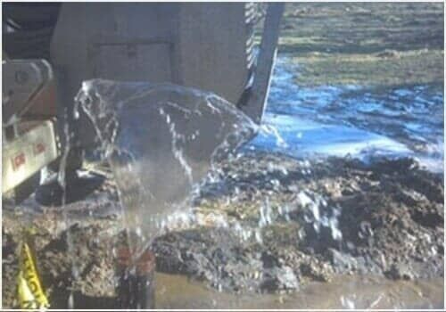 Excavator bucket dumping wet dirt and water into a muddy pile outdoors near snow and ice.