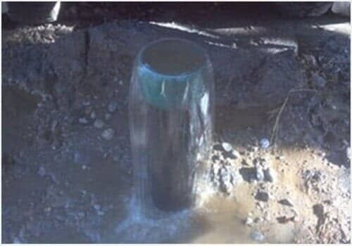 A black cylindrical pipe with a blue rim protrudes from muddy ground in bright sunlight.