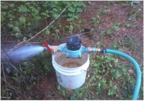 Water sprinkler spraying from a white bucket on grass, connected to a green hose and red valve.