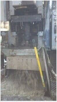 Industrial machine with a yellow-handled broom leaning beside a chute, spilling material onto the floor.
