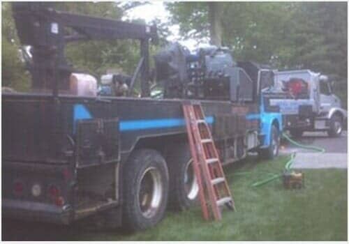 Large blue truck carrying machinery beside another truck, with a ladder leaning on the trailer in a grassy yard.
