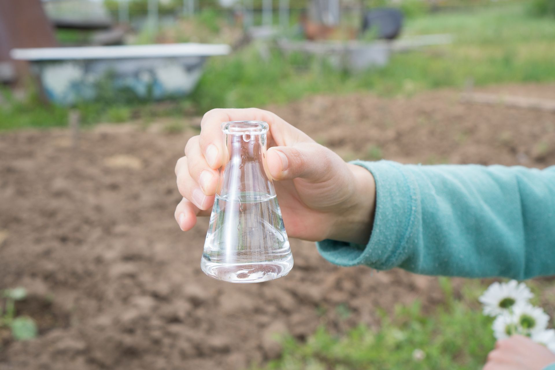 Hand holding a small clear glass flask in a garden bed, with soil and flowers in the background