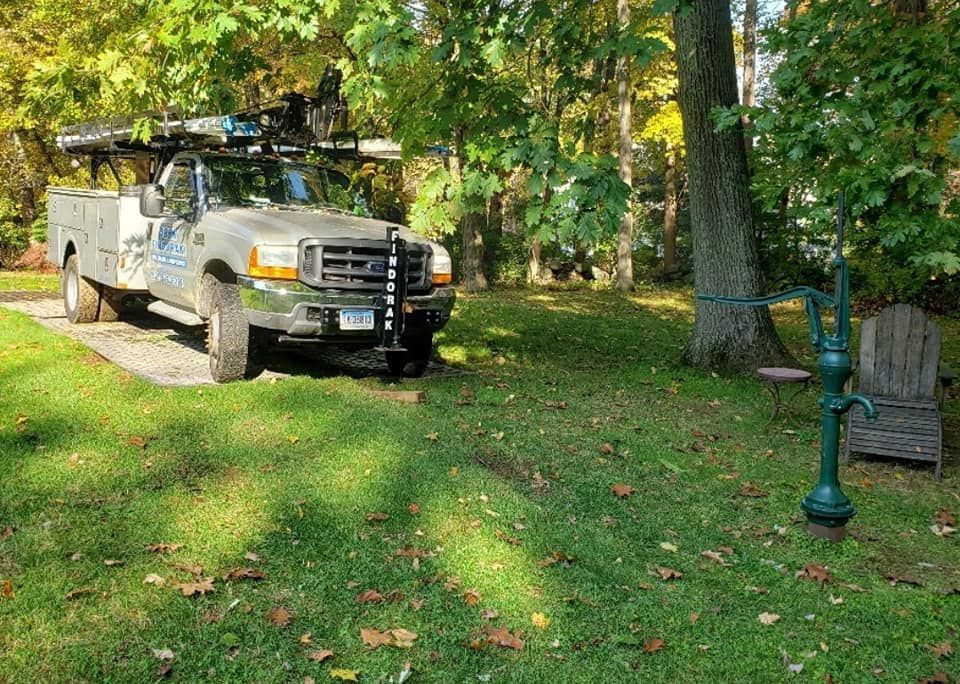 White utility truck parked on a grassy driveway under trees beside a lamp post and chair
