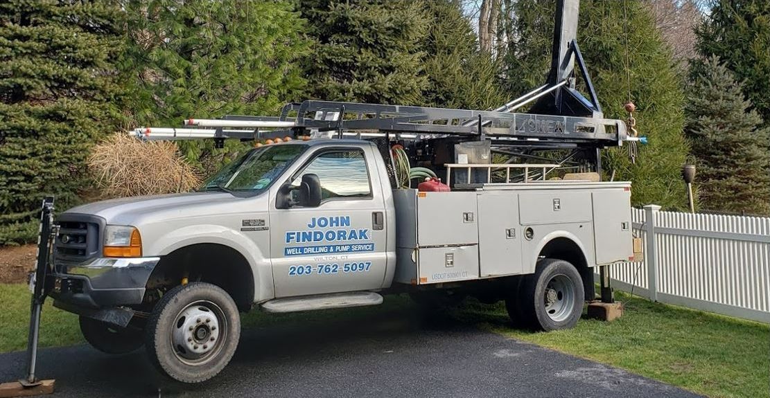 White utility service truck with ladder rack parked on a driveway beside a fence and trees