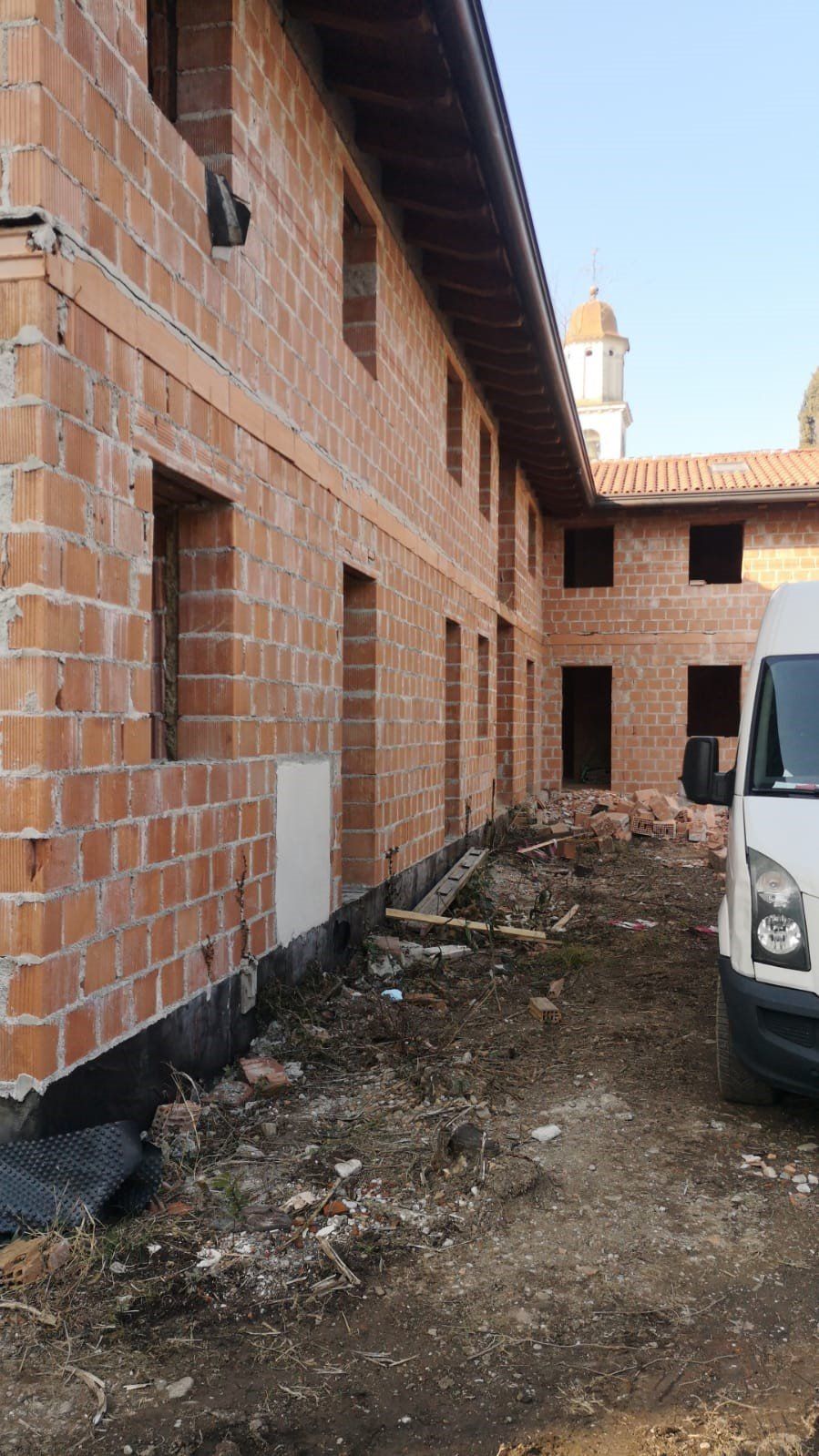 Esterno di un edificio in costruzione con muri in mattoni rossi, finestre e pavimento in terra battuta. Un furgone bianco è parcheggiato nelle vicinanze.