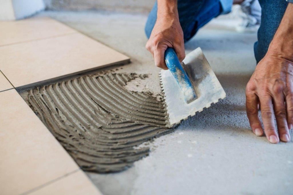 Persona che piastrella un pavimento, utilizzando una cazzuola dentata per stendere l'adesivo sul cemento, con le piastrelle nelle vicinanze.