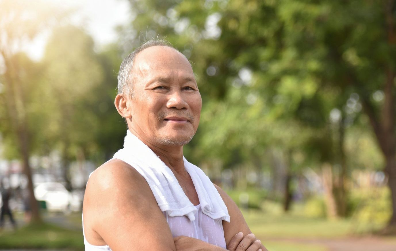 Smiling man with folded arms and a towel over his shoulder in a sunny park