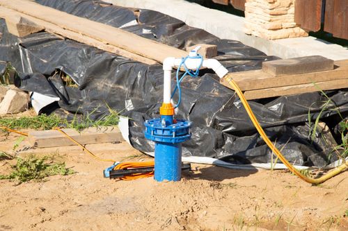 Blue well pump with white pipe and yellow hose on a sandy construction site, near black plastic sheeting.