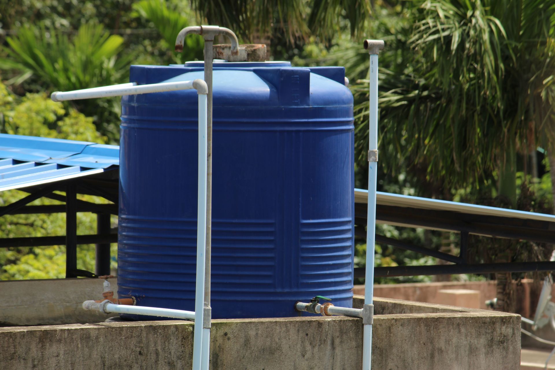 Blue plastic water tank on a concrete platform with metal pipes, outdoors near palm trees.