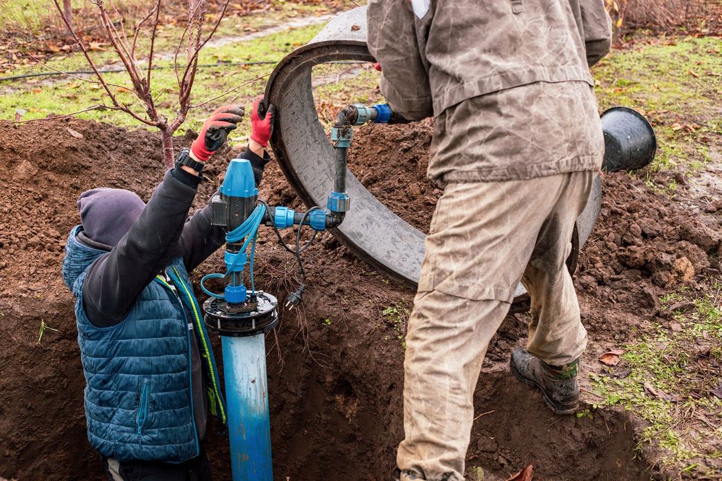 Two workers install blue piping beside a concrete utility tank in muddy ground.