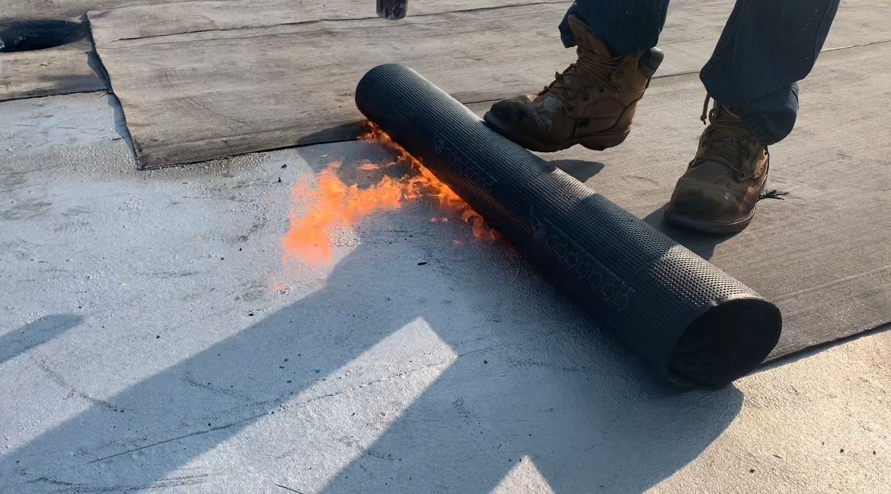 Person using a torch to apply a rolled roofing membrane to a flat roof, with flames visible.