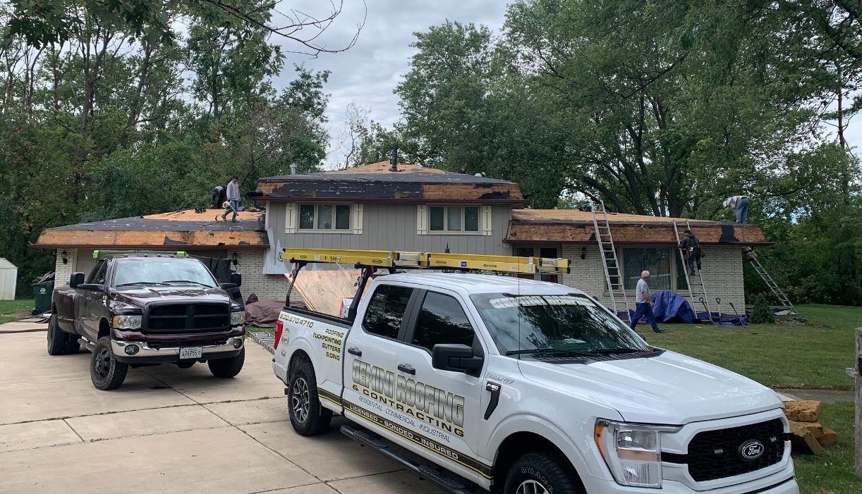 Roofing contractors replacing shingles on a two-story house. Trucks are parked in the driveway; workers are on the roof.
