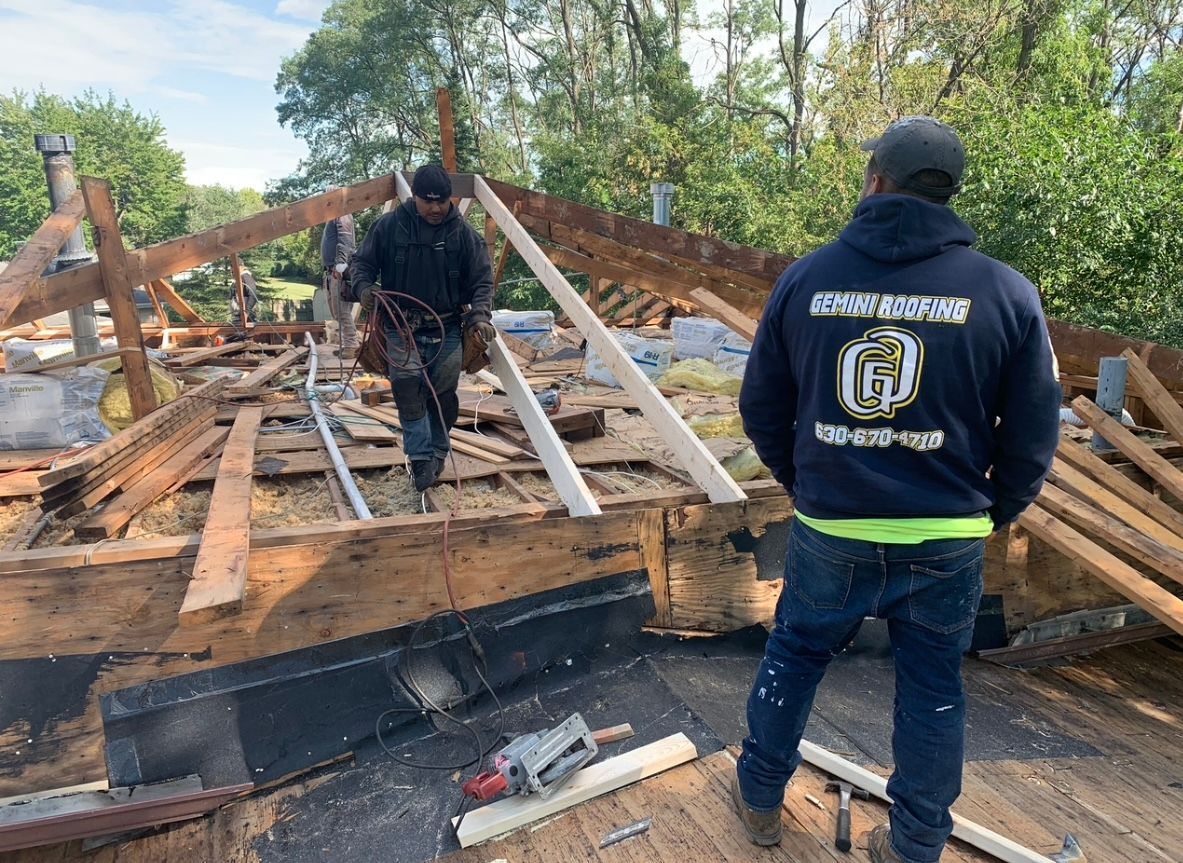 Two roofers working on a partially dismantled roof. One carries tools, and the other wears a blue hoodie with a company logo.