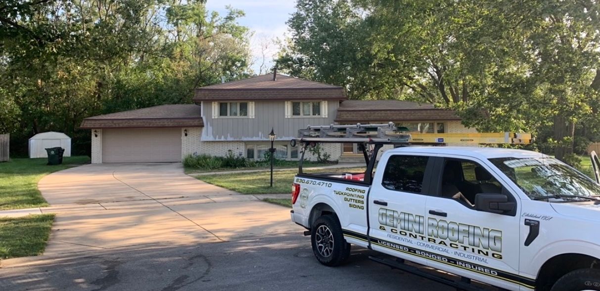 A white pickup truck with company logos parked in front of a two-story house with a gray exterior and a brown roof.
