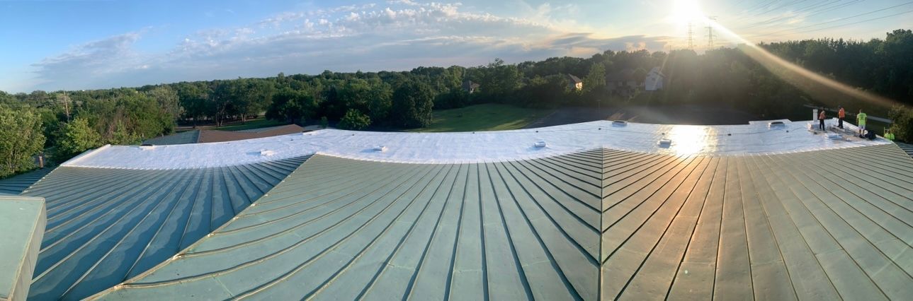 Aerial view of a building with a metal roof, trees, and a bright sky. The sun shines brightly on the right side of the image.