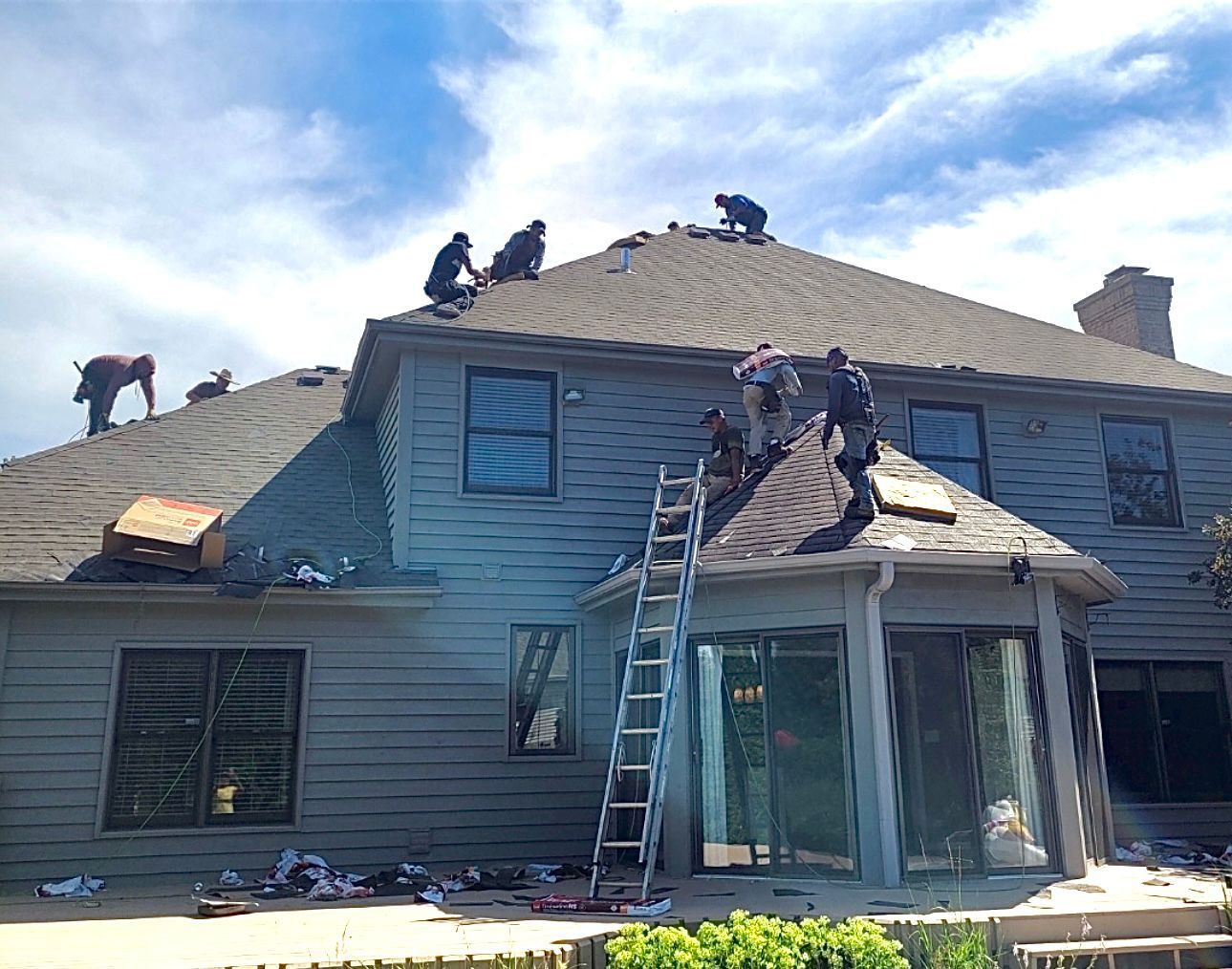 Roofers working on a gray shingled roof of a two-story house on a sunny day. A ladder rests against the roof.