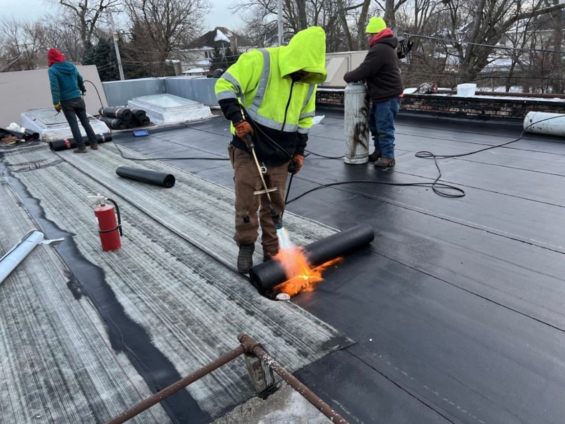 Workers on a flat roof use a torch to heat a black roofing membrane. One worker in yellow is in focus.