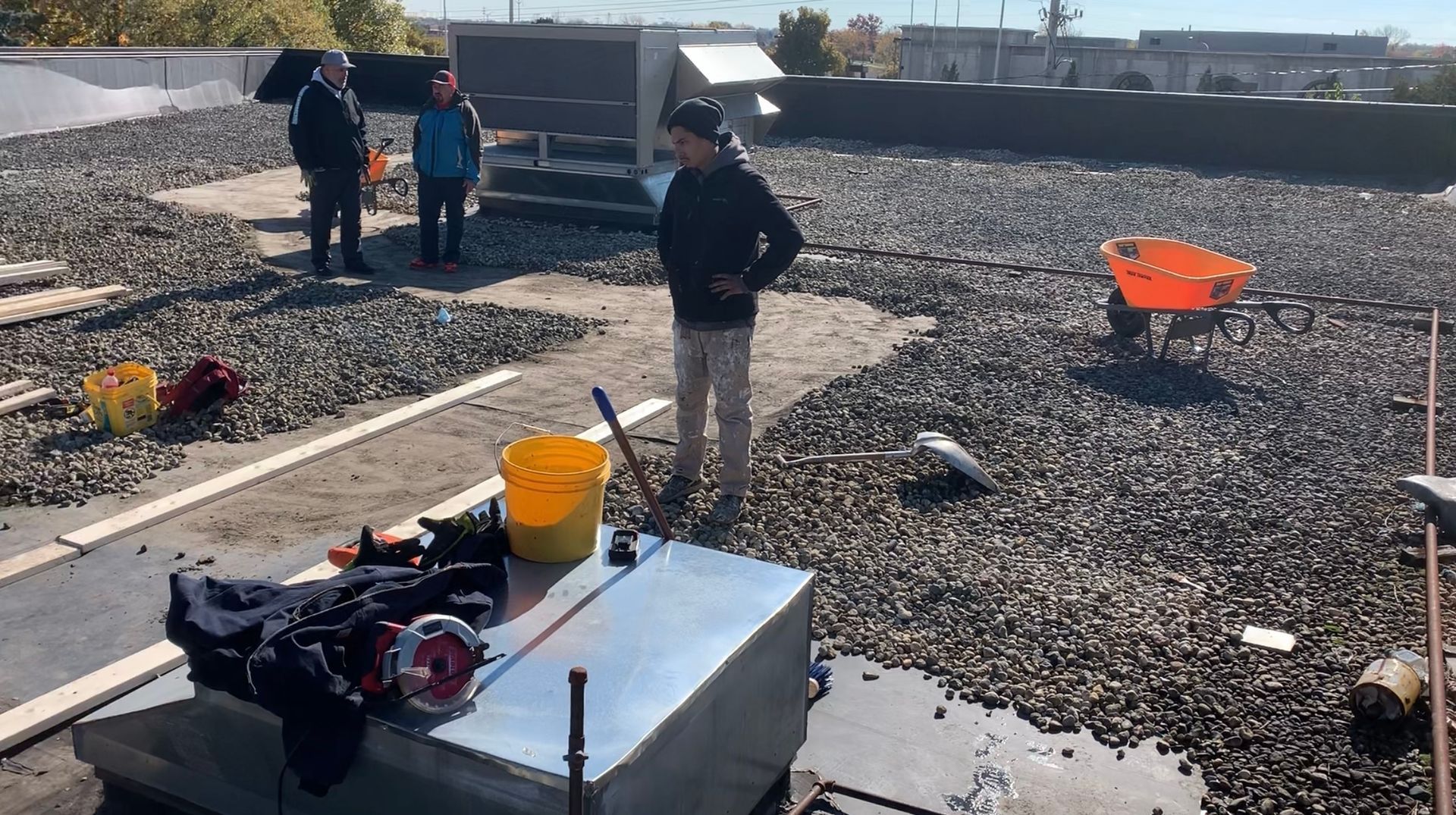 People work on a rooftop covered in gravel. One person stands near a metal box, while others observe.