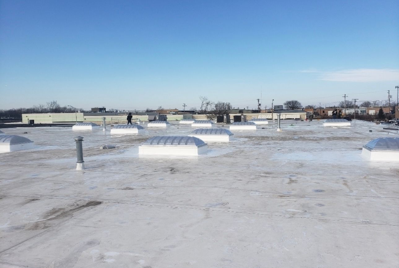 White commercial rooftop with several dome skylights, a clear blue sky, and distant buildings.